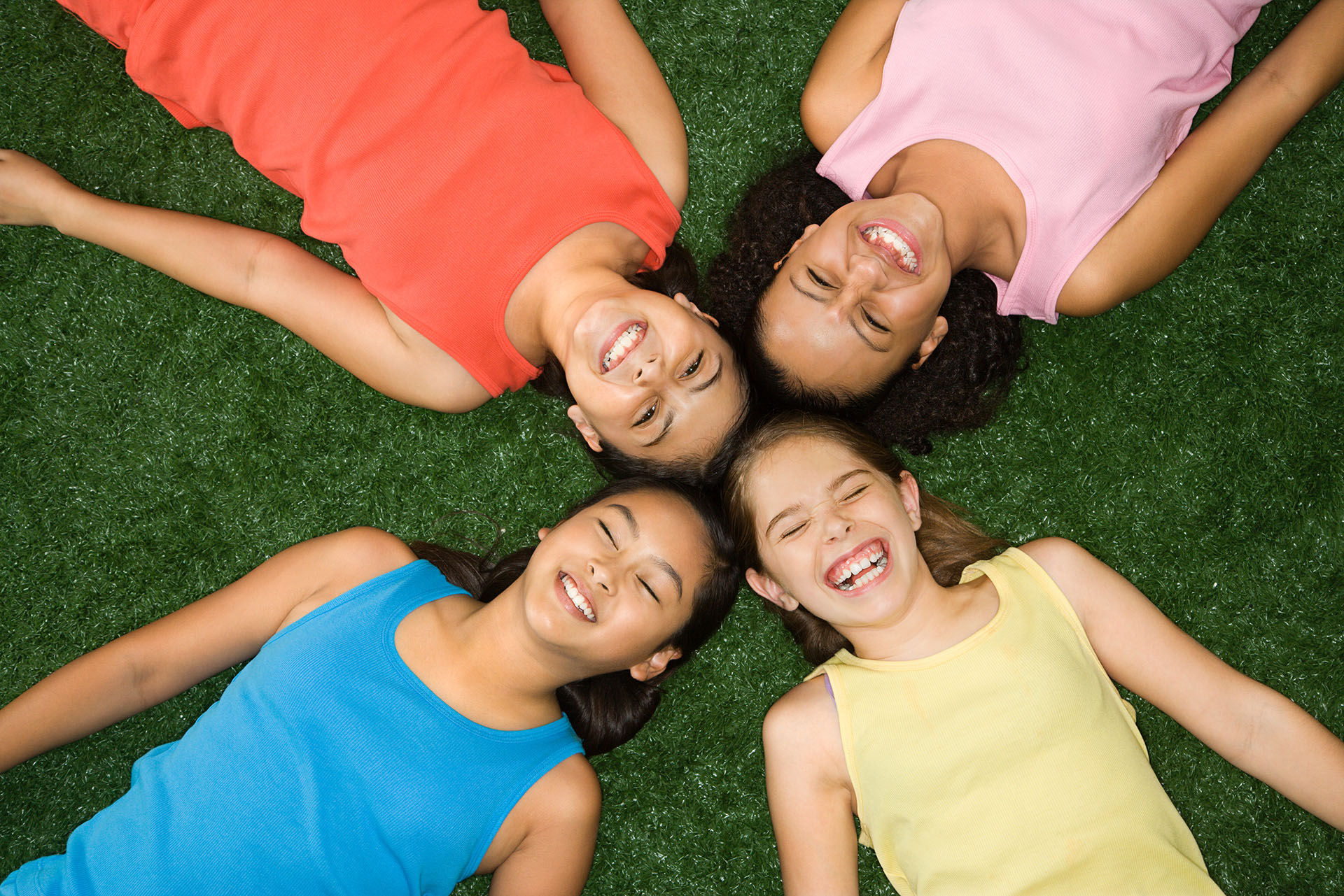 Above view of four girls lying on artificial grass with heads together laughing.